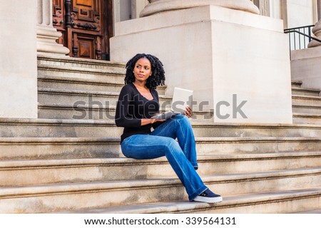 African American college student studying in New York. Wearing long sleeves, V neck top, jeans, leather sneakers, a girl sitting on stairs, working on laptop computer. Filtered look with blue tint.