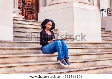African American college student studying in New York. Wearing long sleeves, V neck top, jeans, leather sneakers, a girl sitting on stairs, working on laptop computer. Filtered look with purple tint.