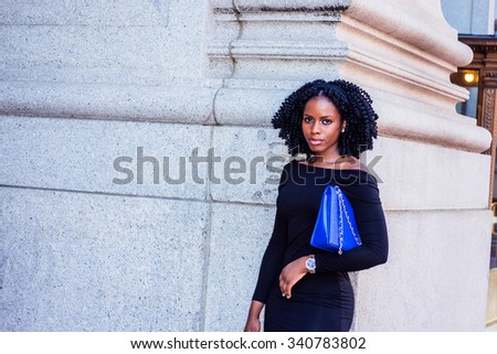 African American Businesswoman working in New York. Wearing long sleeve, slim off shoulder dress, carrying blue bag under arm, a young black lady standing on street. Filtered look with purple tint.