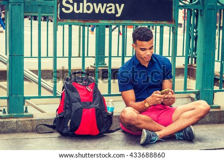 African American college student traveling, studying in New York, wearing blue short sleeve shirt, red shorts, sneakers, bag with bottle water on ground, sitting on street by Subway sign, texting.