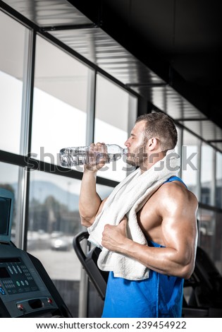 Young adult man drinking bottle of water on treadmill in gym.