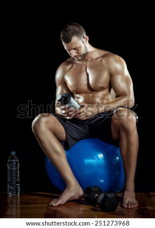 Young adult man drinking protein shake in gym. Black background.