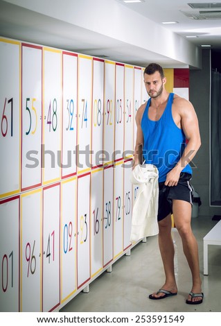 Young adult fit man standing next to lockers in locker room of fitness facility.