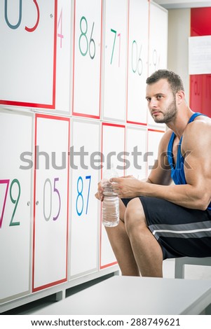 Young adult tired man drinking water in locker room of gym.