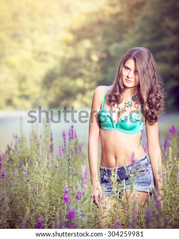 Beautiful young girl standing in high grass and flowers on river bank wearing bra and jeans shorts.