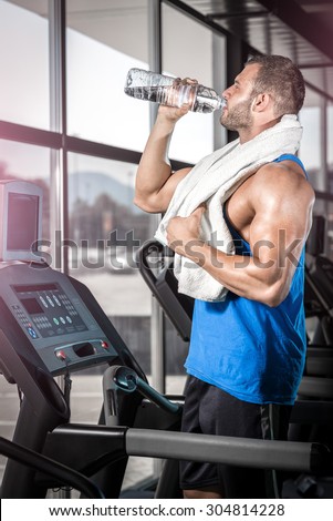 Young adult man drinking bottle of water on threadmill in gym.