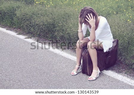 Desperate young girl on a suitcase at the border of a road. Desperation and loneliness concept