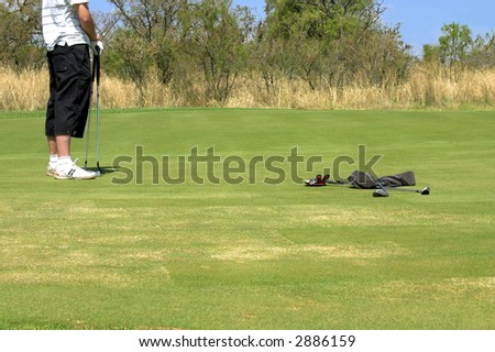 Golfer standing on the green with two golf clubs in his hand. Wet towel and another two golf clubs are lying next to him on the grass. Shot on South African golf course on a sunny day.