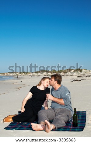 Young married couple sitting on the white sandy beach on their winter vacation in South Africa. They are enjoying the wine under the blue sky.