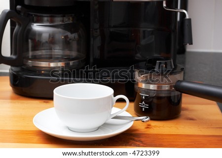Closeup of white cup with coffee machine in the background and pot full of coffee next to it