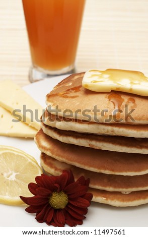 Stack of flapjacks with syrup, slices of lemon, cheese and red flower. Served with a glass of fruit juice