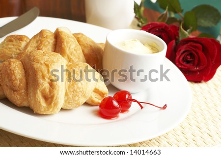 Breakfast plate with croissants, butter and glazed maraschino cherries with red roses in the background