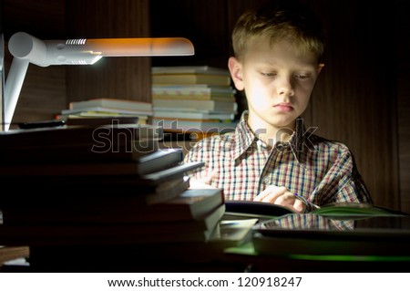 Boy reading a book by the light of a desk lamp.