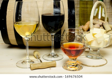 Red and white wine in glasses, brandy. On the background of a barrel and wine corks basket, bowl with cheese and a bottle of wine, a corkscrew.