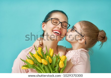 Happy women\'s day! Child daughter is congratulating mom and giving her yellow flowers tulips. Mum and girl smiling and hugging on light blue background. Family holiday and togetherness.