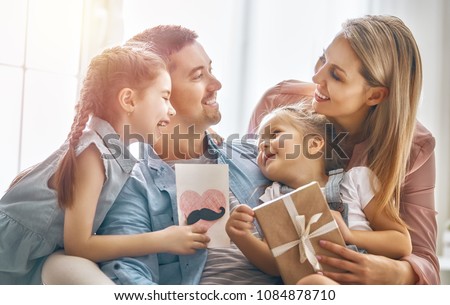 Happy father's day! Children daughters and their mom are congratulating dad and giving him postcard and gift box. Daddy and girls smiling and hugging. Family holiday and togetherness.
