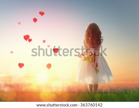 Sweet child girl looking at red balloons. Little child girl holding bouquet of flowers. Balloons in shape of heart flying in the sunset sky. Wedding, Valentine, love concept.