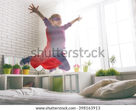 Child girl in Superhero\'s costume plays. The child having fun and jumping on the bed.