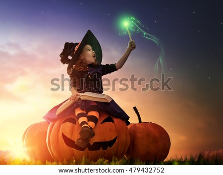 Happy Halloween! Cute cheerful little witch with a magic wand and book of spells. Beautiful child girl in witch costume sitting on the big pumpkin, conjuring and laughing.