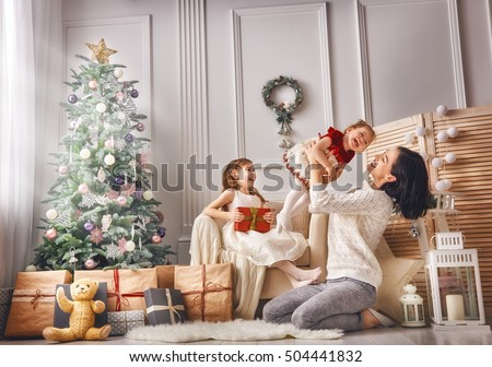 Merry Christmas and Happy Holidays! Cheerful mom and her cute daughters girls exchanging gifts. Parent and two little children having fun and playing together near Christmas tree indoors.