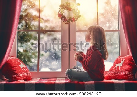 Merry Christmas and happy holidays! Cute little girl sitting by the window with a cup of hot drink and looking at the winter forest. Room decorated on Christmas. Kid enjoys the snowfall.