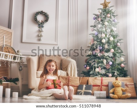 Merry Christmas and Happy Holidays! Cheerful cute little child girl with present. Kid holds a gift box near Christmas tree indoors.