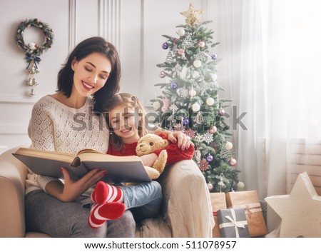 Merry Christmas and Happy Holidays! Pretty young mom reading a book to her cute daughter near Christmas tree indoors.