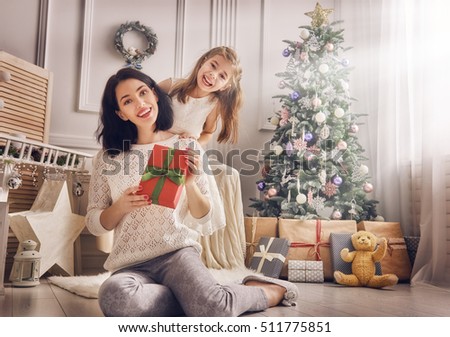 Merry Christmas and Happy Holidays! Cheerful mom and her cute daughter girl exchanging gifts. Parent and little child having fun near Christmas tree indoors. Loving family with presents in room.