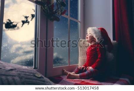 Merry Christmas and happy holidays! Cute little child baby girl sitting by window and looking at Santa Claus flying in his sleigh against moon sky. Room decorated on Christmas. Kid enjoy the holiday.