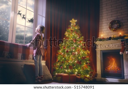 Merry Christmas and happy holidays! Cute little child girl looking by window. Christmas miracle Santa Claus flying in his sleigh against moon sky. Room decorated on Christmas. Kid enjoy the holiday.