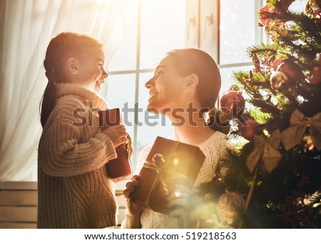 Merry Christmas and Happy Holidays! Cheerful mom and her cute daughter girl exchanging gifts. Parent and little child having fun near Christmas tree indoors. Morning Xmas. Portrait family close up.