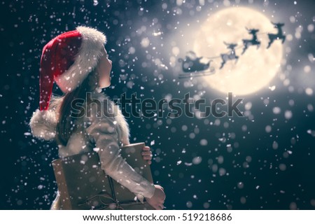 Merry Christmas and happy holidays! Cute little child girl with xmas present. Santa Claus flying in his sleigh against moon sky. Kid enjoy the holiday. Portrait kid with gift on dark background.