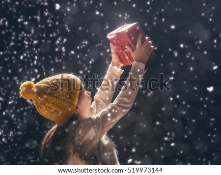 Happy child girl playing on a snowy winter walk. Little girl enjoys the game. Child girl playing outdoors in snow. Outdoor fun for winter vacation. Portrait kid with gift box on dark background.