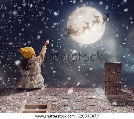 Merry Christmas and happy holidays! Cute little child girl sitting on the roof and looking at Santa Claus flying in his sleigh against moon sky. Kid enjoy the holiday. Christmas legend concept.