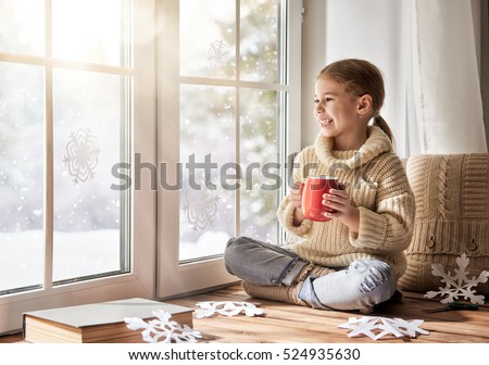 Cute little girl sitting by the window, drinking warm beverage and looking at the winter forest. Child makes paper snowflakes for decoration windows. Kid enjoys the snowfall.