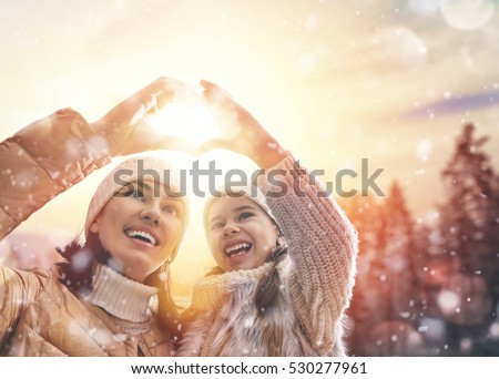 Happy loving family! Mother and child girl having fun, playing and laughing on snowy winter walk in nature. Frost winter season. Mom and kid making hand heart in evening.