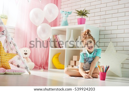 Happy child plays. Little child girl draws with colored pencils indoors. Funny lovely child having fun in children room.