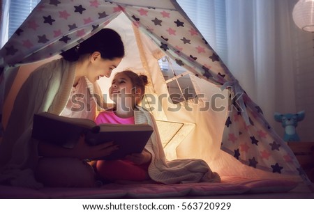 Family bedtime. Mom and child daughter are reading a book with flashlights in tent. Pretty young mother and lovely girl having fun in children room.