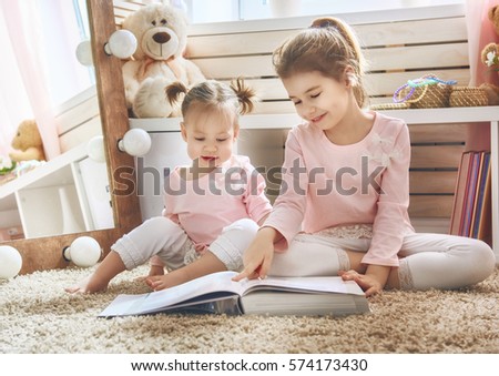 Two cute little children are reading a book. Happy girls play at home. Funny lovely sisters are having fun in children room.