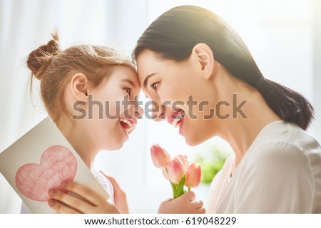 Happy mother\'s day! Child daughter congratulates mom and gives her flowers tulips and postcard. Mum and girl smiling and hugging. Family holiday and togetherness.