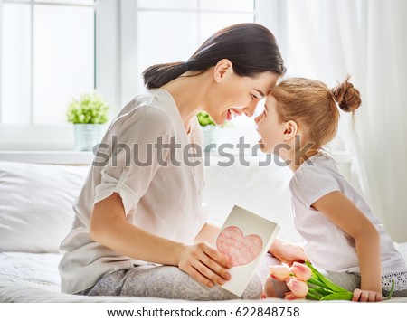 Happy mother\'s day! Child daughter congratulates mom and gives her flowers tulips and postcard. Mum and girl smiling and hugging. Family holiday and togetherness.