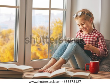 Cute child girl sitting by the window and reading a book in room at home. Beautiful autumn nature.