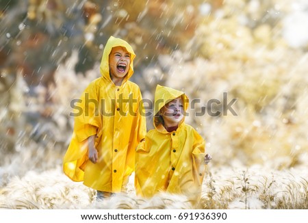 Two happy funny children under the autumn shower. Girls are wearing yellow raincoats and enjoying rainfall. Sisters playing together on the nature outdoors. Family walk in the park.