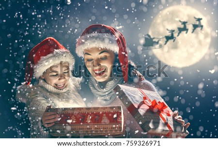 Merry Christmas and Happy Holidays! Mom and daughter are opening gift outdoors. Xmas night. Santa Claus flying in his sleigh against moon sky. Portrait loving family close up.