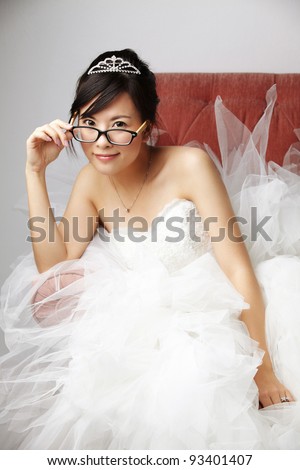 Young beautiful Asian Bride wearing bridal tiara in white bride dress with smile wearing eye glasses look at the camera sitting on a pink armchair under studio light