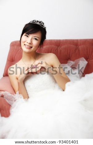 Young beautiful Asian Bride wearing bridal tiara in white bride dress with smile look at the camera sitting on a pink armchair under studio light