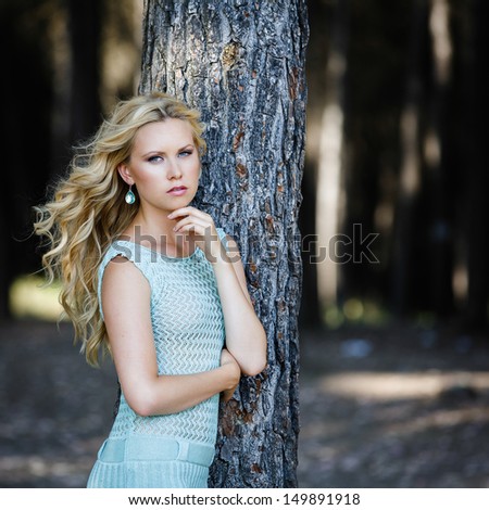 Beautiful girl with blonde hair and blue eyes, leaning against a tree in the forest in a sunny day