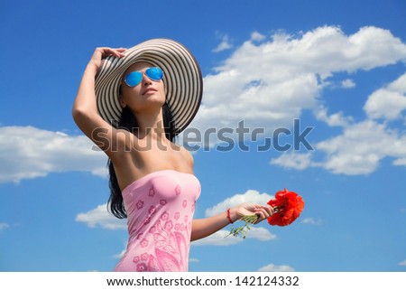Portrait of a sexy woman in the big stripy hat with the poppy flowers in her hand, cloudy sky background