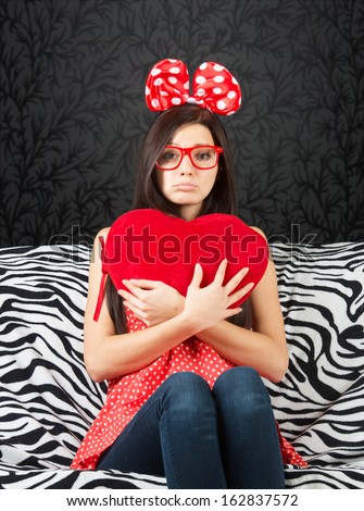 Sad girl with a big hair bow and a heart cushion sitting on the sofa and looking at camera