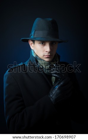 Portrait of a young man in the black trench coat and hat standing in the darkness